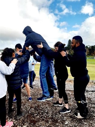 A boy in a hooded sweatshirt stands on a low wire rope supported by four other teenagers.