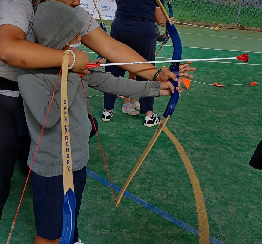 Ākonga is standing with a bow and arrow in position learning how to shoot at the Halberg Sports Day. An adult is standing behind the ākonga helping hold the equipment.