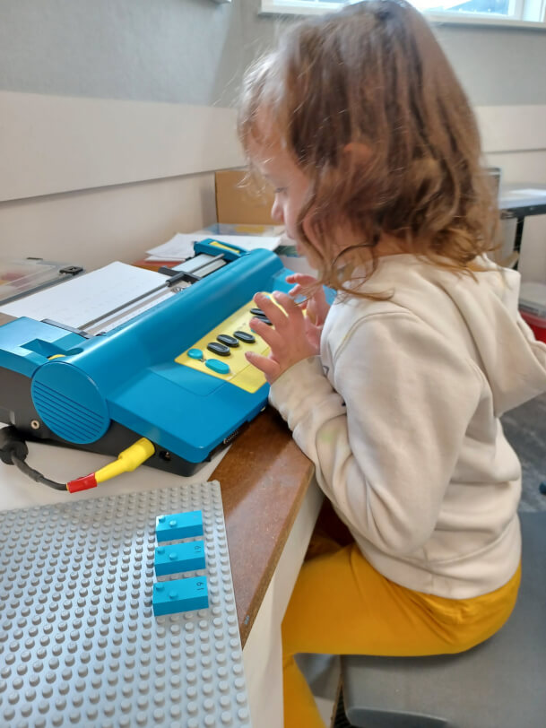 Child is learning the letter ‘i’. LEGO Braille Bricks are set up on a baseboard and she is brailling these on the Mountbatten.