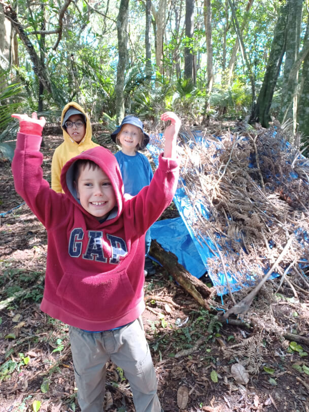Boy in red smiles with hands in the air, stood in front of his 2 smiling friends and a tarpaulin covered in sticks and leaves.